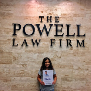 A smiling young girl stands in front of "The Powell Law Firm" wall sign, holding a promotional poster for "Paul" with the slogan "More Lawyer, Less Fee," representing a personal injury law firm in Las Vegas.