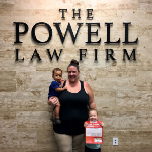 A woman standing with two young children in front of "The Powell Law Firm" sign, possibly a satisfied client of the Las Vegas personal injury lawyers.