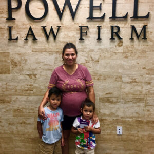 A smiling woman with two young children stands in front of "The Powell Law Firm" sign, representing personal injury legal support in Las Vegas.
