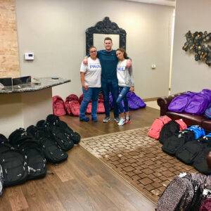 Three smiling individuals stand surrounded by colorful backpacks in a Las Vegas law firm, showing their dedication to community support and personal injury advocacy.