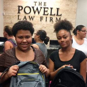Two smiling young women stand in front of "The Powell Law Firm" sign in Las Vegas, holding backpacks, highlighting the firm's dedication to community support and personal connections beyond personal injury cases.