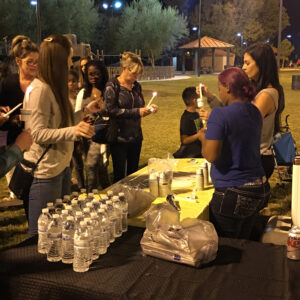 Outdoor night scene shows a group gathered, lighting candles at a table offering water bottles; symbolizing support for personal injury victims in Las Vegas.