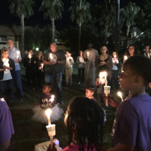 A diverse group of people, including children, hold candles in a vigil at night, possibly in Las Vegas, demonstrating community support and solidarity, suggesting themes of remembrance and support for those affected by personal injury or loss.
