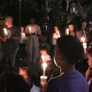 A diverse group of people, including adults and children, gather outdoors at night holding lit candles at a vigil in Las Vegas, possibly for those affected by personal injury or tragedy, with trees and buildings in the background.