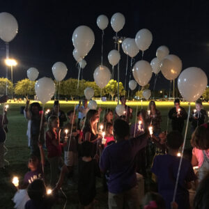 Outdoor night scene of a diverse group of people, including children, holding candles and white balloons at a vigil, possibly related to personal injury victims, reflecting community support and healing.