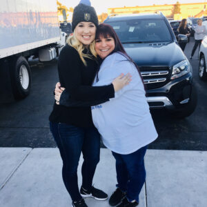 Two smiling women hugging in an outdoor parking lot setting, near a truck and a luxury car, possibly representing personal injury support or community involvement in Las Vegas.