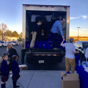 A team unloads blue tote bags from a truck in Las Vegas, while children in school uniforms and adults observe, suggesting a charitable event or community outreach supported by the personal injury law firm.