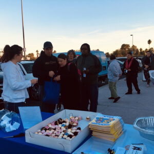A group of people are gathered around a table covered in a blue tablecloth at an outdoor community event, with cupcakes, informational pamphlets and giveaway entry forms on the table, likely hosted by a Las Vegas personal injury law firm.