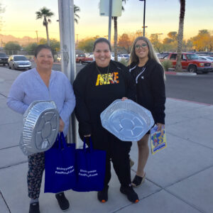 Three smiling women holding foil trays and 'Giving Thanks PaulPowell.com' bags outside in Las Vegas at sunset, likely representing community outreach or volunteer work by a personal injury law firm.