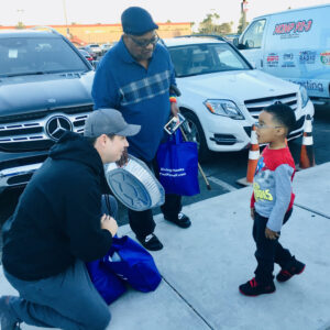 Image shows a law firm representative kneeling to speak with a young boy, while an older man stands next to them holding a bag that says "Giving Thanks PaulPowell.com"; the scene is set in a parking lot in Las Vegas, suggesting the firm's involvement in community service and personal injury representation.