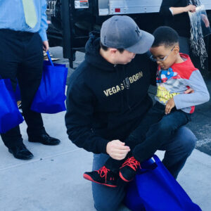 A volunteer from a Las Vegas personal injury law firm kneels to comfort a young boy, handing out blue bags, while others unload a truck in the background, signifying community outreach and support.