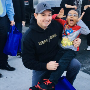 A smiling lawyer wearing a "Vegas Born" hoodie kneels holding a joyful child in his arms at a community event, symbolizing the firm's commitment to Las Vegas and dedication to representing personal injury clients with care.