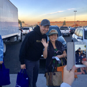 Smiling man with a "Vegas" sweatshirt and an Asian woman posing and waving while someone takes their picture in a parking lot with cars and a large truck, possibly related to a personal injury support event.