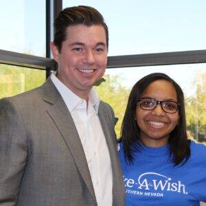 A smiling lawyer in a suit stands next to a smiling woman in a Make-A-Wish shirt, symbolizing community involvement and support for charitable causes by the Las Vegas personal injury law firm.