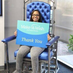 A smiling young woman sits in a unique metal chair holding a "Make-A-Wish Southern Nevada Thank You" sign, symbolizing the Las Vegas law firm's commitment to charitable giving and supporting personal injury victims through community involvement.