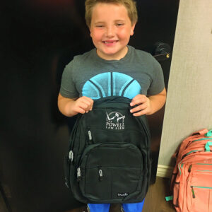 A smiling young boy stands holding a black backpack emblazoned with The Powell Law Firm logo, suggesting the firm's involvement in community support and potentially hinting at cases involving children's well-being or accidents.