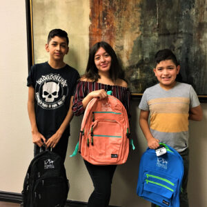 Three children pose with colorful backpacks bearing the logo of a Las Vegas personal injury law firm, The Powell Law Firm, showcasing their commitment to community support.