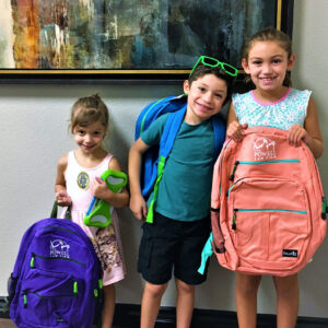 Three smiling children stand in front of an abstract painting, each holding or wearing a brightly colored backpack with the Powell Law Firm logo, promoting a family-friendly image for the Las Vegas personal injury law firm.