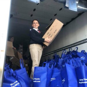 A young volunteer in a school uniform loads boxes of turkeys and blue bags labeled "Giving Thanks Paul Powell" into a truck, representing the Las Vegas personal injury law firm's commitment to community service and helping those in need during the holiday season.