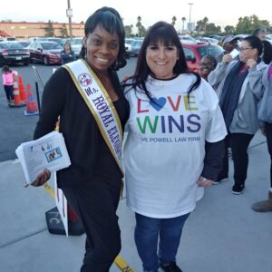 Two women smiling, one wearing a "Ms. Royal Elegance" sash and holding YMCA pamphlets, the other in a "Love Wins, The Powell Law Firm" t-shirt, at a community event in Las Vegas.