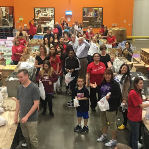 A large group of diverse volunteers pack bags of food in a warehouse, showing a Las Vegas personal injury law firm's commitment to community support and care for those in need, emphasizing their dedication to helping people.