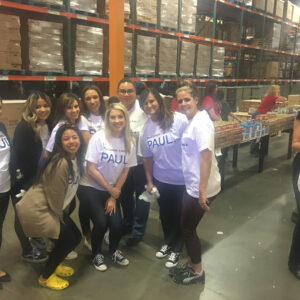 A group of employees from a Las Vegas personal injury law firm wearing matching t-shirts pose for a photo in a large warehouse filled with food donations, showcasing their commitment to community service.