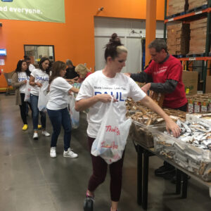A team of volunteers wearing "More Lawyer. Less Fee. PAUL" t-shirts pack food items in a warehouse, showcasing community involvement by the Las Vegas personal injury law firm.