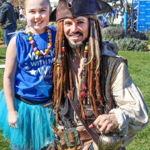 A girl in a blue tutu smiles next to a man dressed as Jack Sparrow at a Make-A-Wish Foundation event in Las Vegas, showcasing community support from a Personal Injury Law Firm.