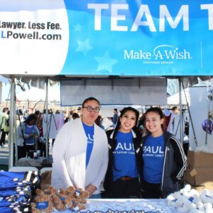 Image shows three women wearing "Paul Powell" T-shirts at the Make-A-Wish Foundation's "Walk for Wishes" event, representing the Las Vegas personal injury law firm, Paul Powell, with a banner that reads "More Lawyer. Less Fee."