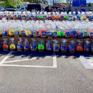A large collection of colorful Easter baskets filled with treats and wrapped in clear plastic wrap are displayed on tables in a parking lot, with people milling around in the background.