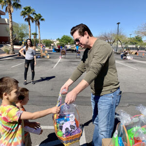 Outdoors event where a man hands a basket to two children, with other people and baskets nearby in a sunny parking lot, potentially symbolizing personal care and community support after an injury.