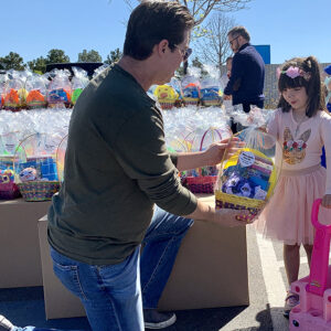 Man kneels to present an Easter basket to a child during a community event hosted by a Las Vegas personal injury law firm.