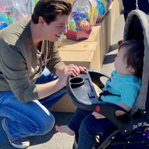 Las Vegas attorney kneels down and smiles at a baby in a stroller during a community event with Easter baskets, representing the firm's dedication to families and personal injury cases.