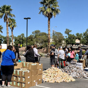 A Las Vegas personal injury law firm hosts a community outreach event distributing food and beverages to a diverse group of people in a parking lot under a bright blue sky with palm trees.