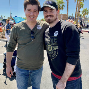 Two Las Vegas personal injury lawyers, smiling and posing for a photo at a community event, wearing "More Lawyer, Less Fine" buttons, with palm trees and the American flag in the background.