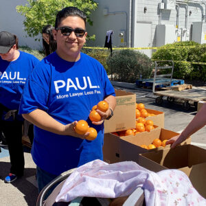 A smiling man in a blue "PAUL. More Lawyer, Less Fee." t-shirt holds oranges at a community outreach event for a Las Vegas personal injury law firm, with boxes of oranges and people nearby.