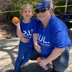 A smiling woman in a "PAUL. Lawyer. Less Fee." t-shirt and hat kneels next to a young girl, both holding oranges, symbolizing support and community involvement related to injury law in Las Vegas.