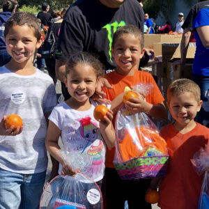A group of smiling children hold oranges and gift bags with "More Lawyer. Less Fee." stickers at a community event supported by a Las Vegas personal injury law firm.
