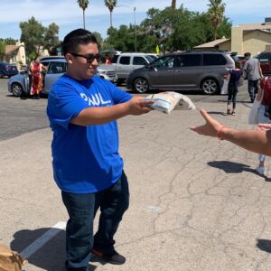 A volunteer hands a bag of goods to a person in a parking lot in Las Vegas, with palm trees and cars in the background, showcasing the law firm's community support.