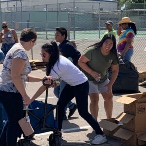A diverse group of volunteers in Las Vegas, possibly associated with a personal injury law firm, work together at a food drive, pulling wagons filled with boxes and canned goods near a tennis court fence, demonstrating community involvement and support.