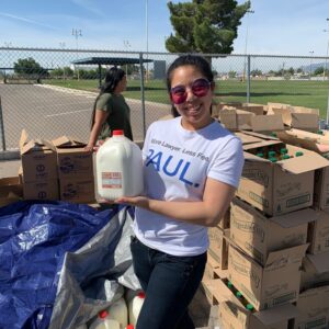 A woman in Las Vegas wearing a "More Lawyer, Less Fee" t-shirt stands smiling while holding a gallon of milk, surrounded by boxes of milk and vegetable oil for a community outreach event, showcasing the firm's commitment to service beyond personal injury law.
