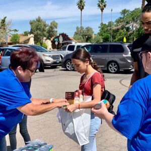 A group of volunteers in blue shirts are assisting a young girl by placing food items into a white bag outdoors, with cars and palm trees visible in the background in Las Vegas.