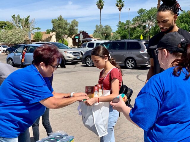 A group of volunteers in blue shirts are assisting a young girl by placing food items into a white bag outdoors, with cars and palm trees visible in the background in Las Vegas.