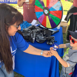 A Las Vegas Law Firm representative hands a small prize to a child at a community event with a prize wheel and branded items.