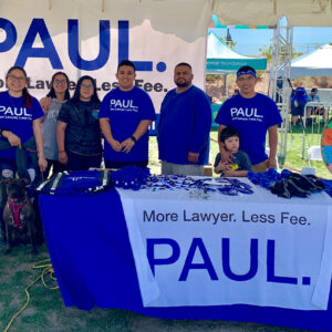 A group of people wearing blue shirts with the logo "PAUL More Lawyer. Less Fee." stand behind a table with the same logo at an outdoor event, alongside a spin wheel and a dog, promoting their law firm specializing in personal injury cases in Las Vegas.