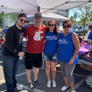 A group of people in "Paul. More Lawyer. Less Fee." shirts pose together in an outdoor setting with tables of backpacks; related to personal injury and community support.