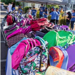 Table filled with colorful backpacks at a Law Firm's community event in Las Vegas, with people and a tent in the background.