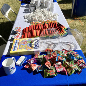 Promotional table displaying giveaway items including branded mugs, wristbands, children's safety books, rainbow-themed items, and Ring Pop candies, representing a personal injury law firm's community engagement in Las Vegas.