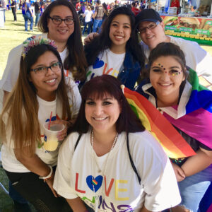A group of diverse women smile in a sunny outdoor setting at a Pride event, wearing "LOVE WINS" t-shirts with the "The Pozoins Law Firm" logo printed on the bottom, and a rainbow flag, showcasing the firm's support for the LGBTQ+ community and commitment to inclusivity, which is relevant for personal injury legal representation.