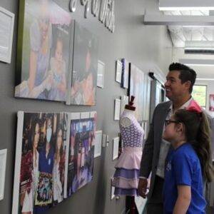 A lawyer and a young girl look at a wall display of photos at a "Journey of a Wish" event, representing community involvement and commitment to children's causes, aligning with a law firm's dedication to justice and support for personal injury victims in Las Vegas.